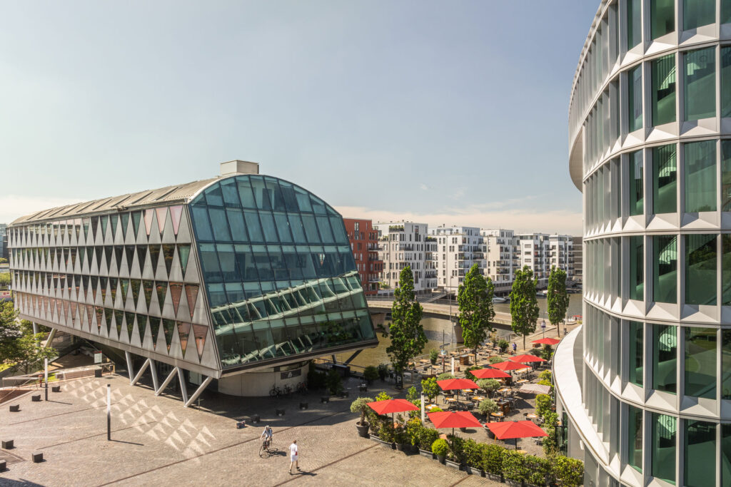 Blick auf den Westhafenplatz vor dem Westhafen Haus mit Außenbereich des Restaurants L'Osteria und Westhafen Brückengebäude